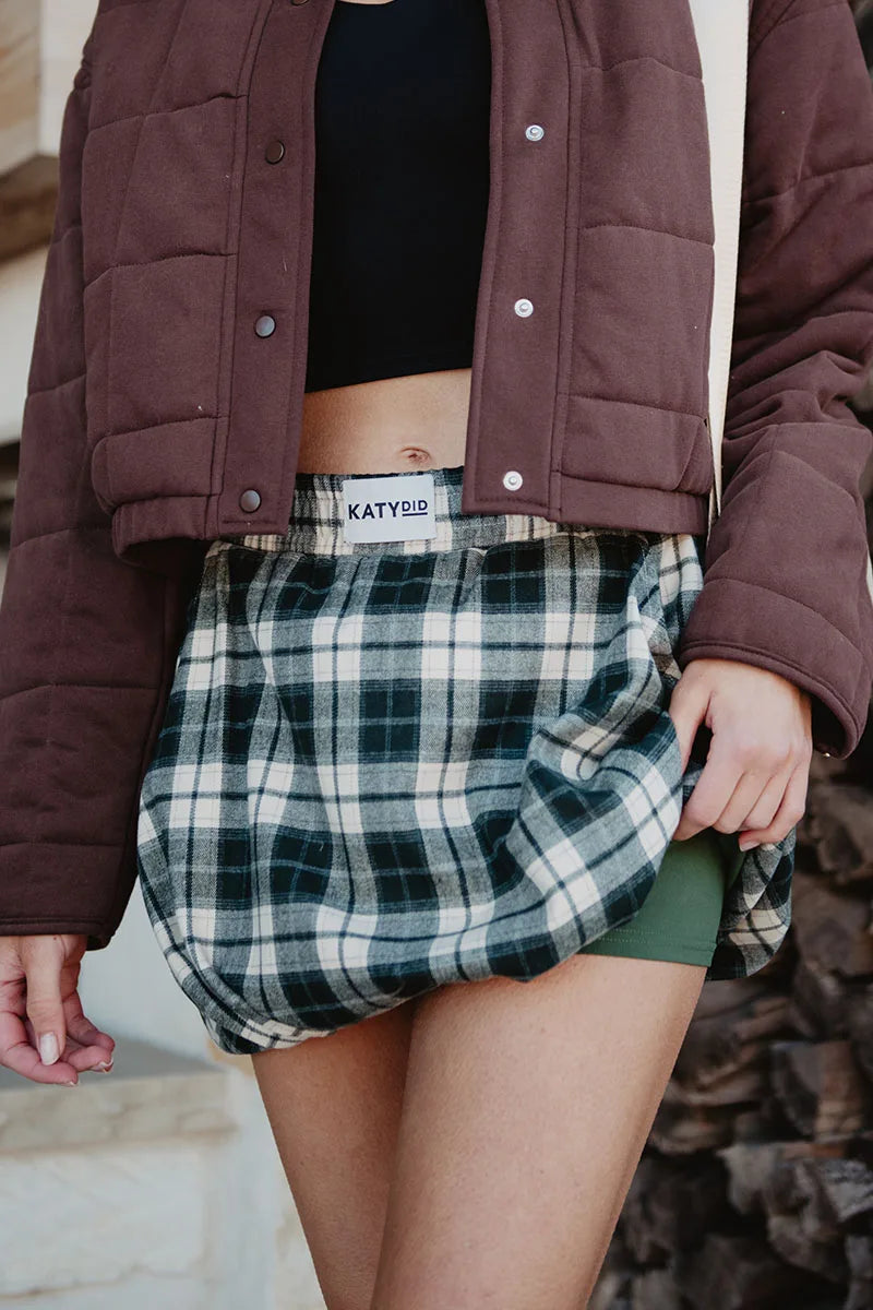 A person wears a brown quilted jacket, black crop top, and a Katydid Green Autumn Plaid Skort. They gently hold the skort to the side, revealing green shorts underneath for added comfort.