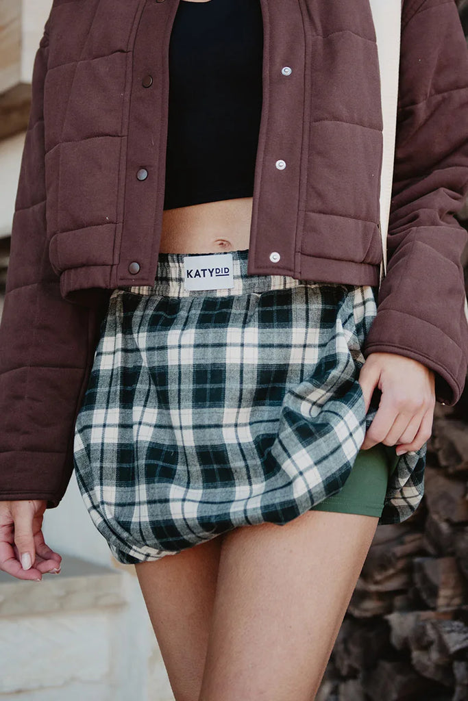 A person wears a brown quilted jacket, black crop top, and a Katydid Green Autumn Plaid Skort. They gently hold the skort to the side, revealing green shorts underneath for added comfort.