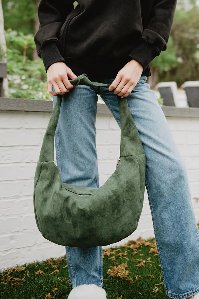 A woman outside stands on grass near a white brick wall, holding the Katydid Green Suede Tote Bag For Women in both hands while wearing a black sweatshirt and blue jeans.