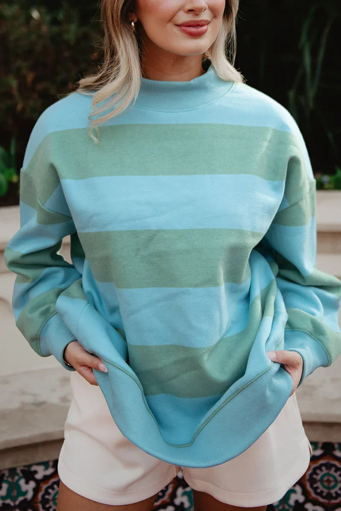 A woman stands outdoors in a Katydid Aqua and Green Striped Mockneck Sweatshirt for women and white shorts, holding the sides of her cozy top. She smiles slightly, with her face partially visible.