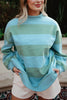 A woman stands outdoors in a Katydid Aqua and Green Striped Mockneck Sweatshirt for women and white shorts, holding the sides of her cozy top. She smiles slightly, with her face partially visible.