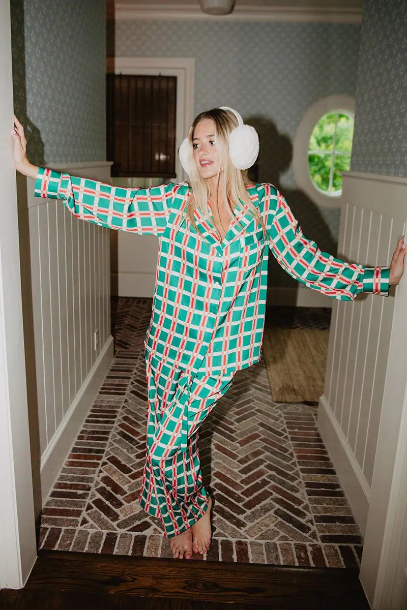 A woman in the Katydid Green  Pink Plaid Pajama Pants Set and white earmuffs leans against a doorway with arms outstretched, standing barefoot on a patterned brick hallway floor.