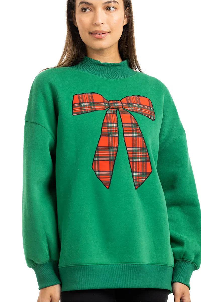 A woman with long brown hair wears the Katydid Red Plaid Bow Mockneck Sweatshirt for women, featuring a large festive bow graphic—a cozy holiday outfit choice—standing against a plain white background.