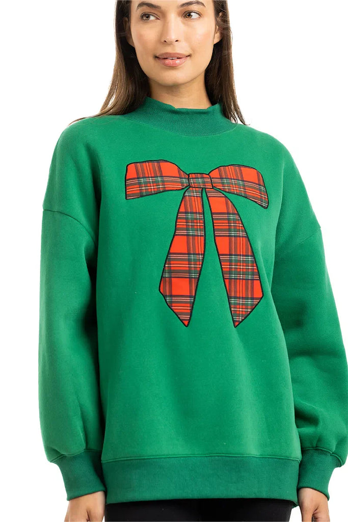 A woman with long brown hair wears the Katydid Red Plaid Bow Mockneck Sweatshirt for women, featuring a large festive bow graphic—a cozy holiday outfit choice—standing against a plain white background.