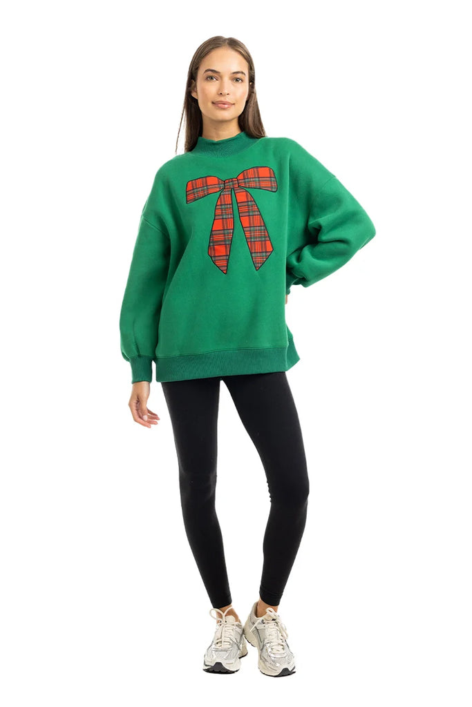 A woman poses against a white background, wearing the Katydid Red Plaid Bow Mockneck Sweatshirt for women—featuring a large plaid bow graphic—paired with black leggings and white sneakers.