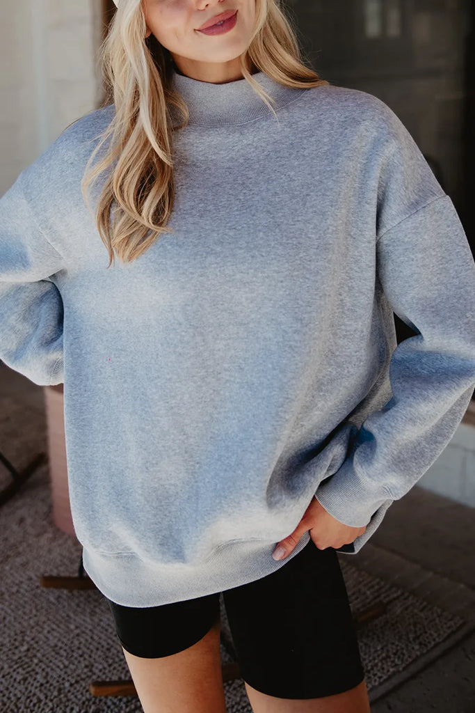 A person with long blonde hair poses casually indoors, wearing the Katydid Heather Essential Mockneck Sweatshirt in cozy heather gray and black shorts, with one hand in their pocket.