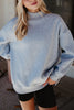 A person with long blonde hair poses casually indoors, wearing the Katydid Heather Essential Mockneck Sweatshirt in cozy heather gray and black shorts, with one hand in their pocket.