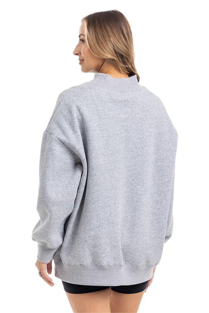 A woman with long, light brown hair wears the Katydid Heather Essential Mockneck Sweatshirt in heather gray, paired with black shorts. She stands against a white background—showcasing this cozy pullover perfect for fall.