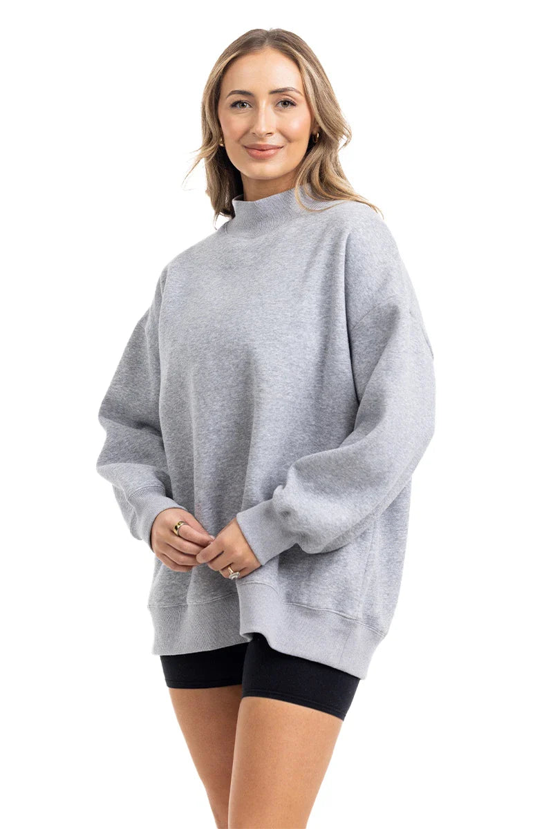 A person with light brown hair is smiling and standing against a white background, wearing the Katydid Heather Essential Mockneck Sweatshirt in heather gray paired with black shorts.