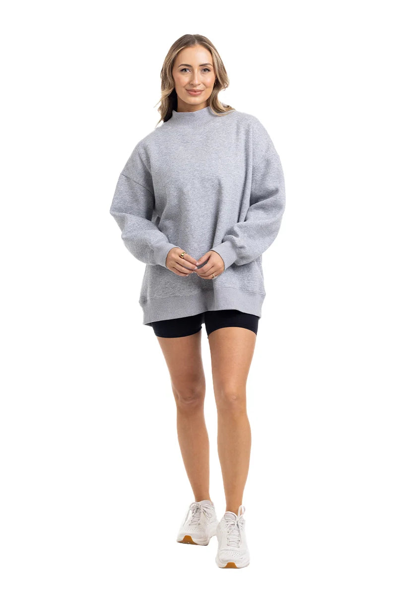 A woman smiles in a Katydid Heather Essential Mockneck Sweatshirt, black shorts, and white sneakers against a white background—a cozy pullover ideal for your fall wardrobe.