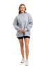 A woman smiles in a Katydid Heather Essential Mockneck Sweatshirt, black shorts, and white sneakers against a white background—a cozy pullover ideal for your fall wardrobe.