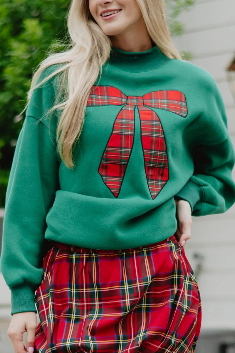A woman wears the Katydid Red Plaid Bow Mockneck Sweatshirt, styled with a matching red plaid skirt for a festive holiday look as she stands outdoors.