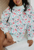 A person wearing the Katydid Holiday Champagne, Coupes  Holly Mockneck Sweatshirt—featuring a colorful wine glass, bottle, and holly design—sits on a white surface with one arm resting on their knee.