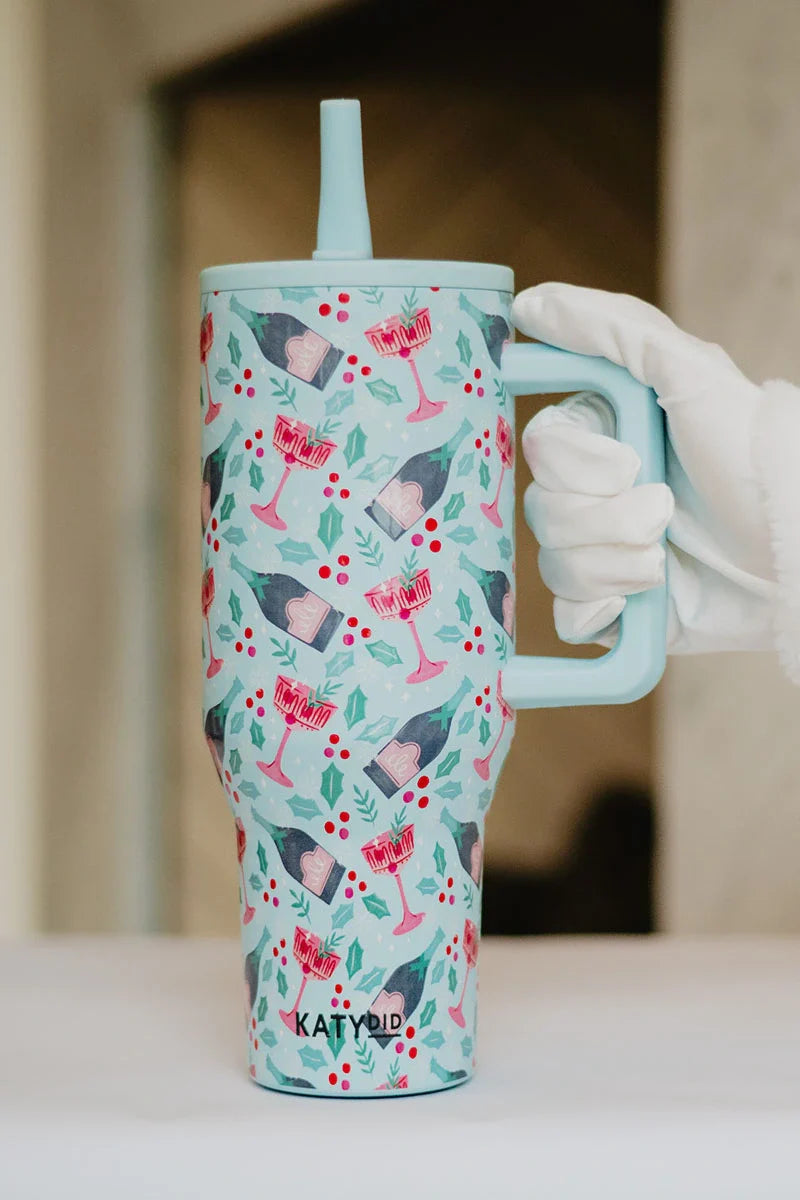 A gloved hand holds the Katydid Holiday Champagne Cheerful Design Flip Top Tumbler, a spill-proof travel cup with handle and straw, decorated with cocktails, books, holly berries, leaves, and KATYDID printed near the base.