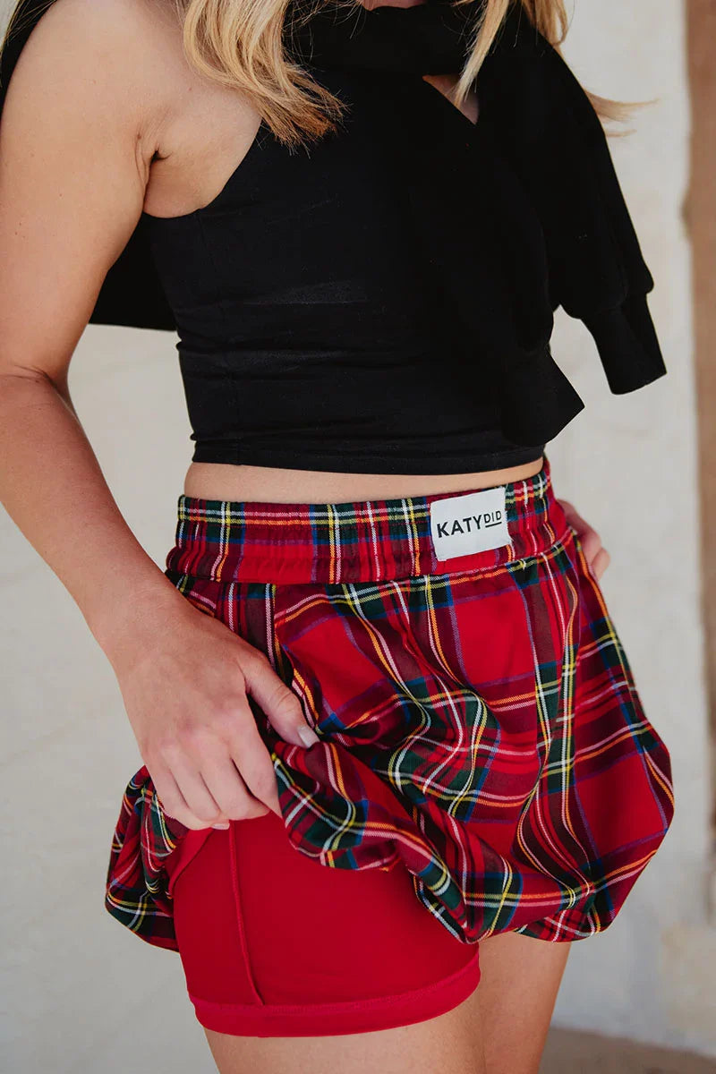 A person in a black sleeveless top and Katydid’s Royal Stewart Plaid Mini Bubble Skirt lifts the skirt to show red liner shorts underneath. The visible torso and upper legs make this a fun, festive holiday party look.