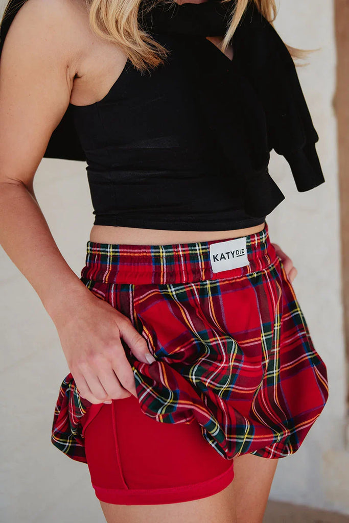 A person in a black sleeveless top and Katydid’s Royal Stewart Plaid Mini Bubble Skirt lifts the skirt to show red liner shorts underneath. The visible torso and upper legs make this a fun, festive holiday party look.