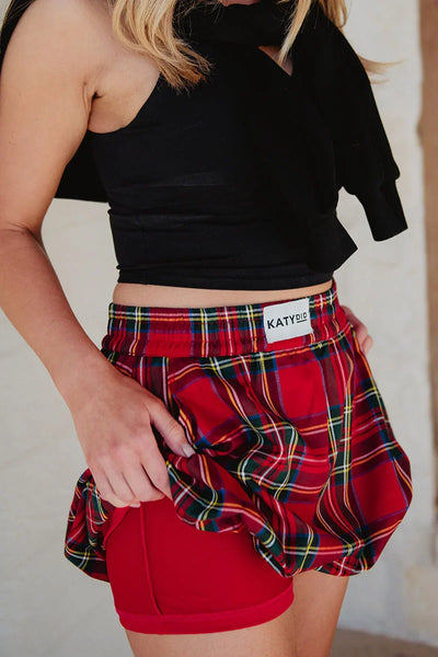 A person in a black sleeveless top and Katydid’s Royal Stewart Plaid Mini Bubble Skirt lifts the skirt to show red liner shorts underneath. The visible torso and upper legs make this a fun, festive holiday party look.