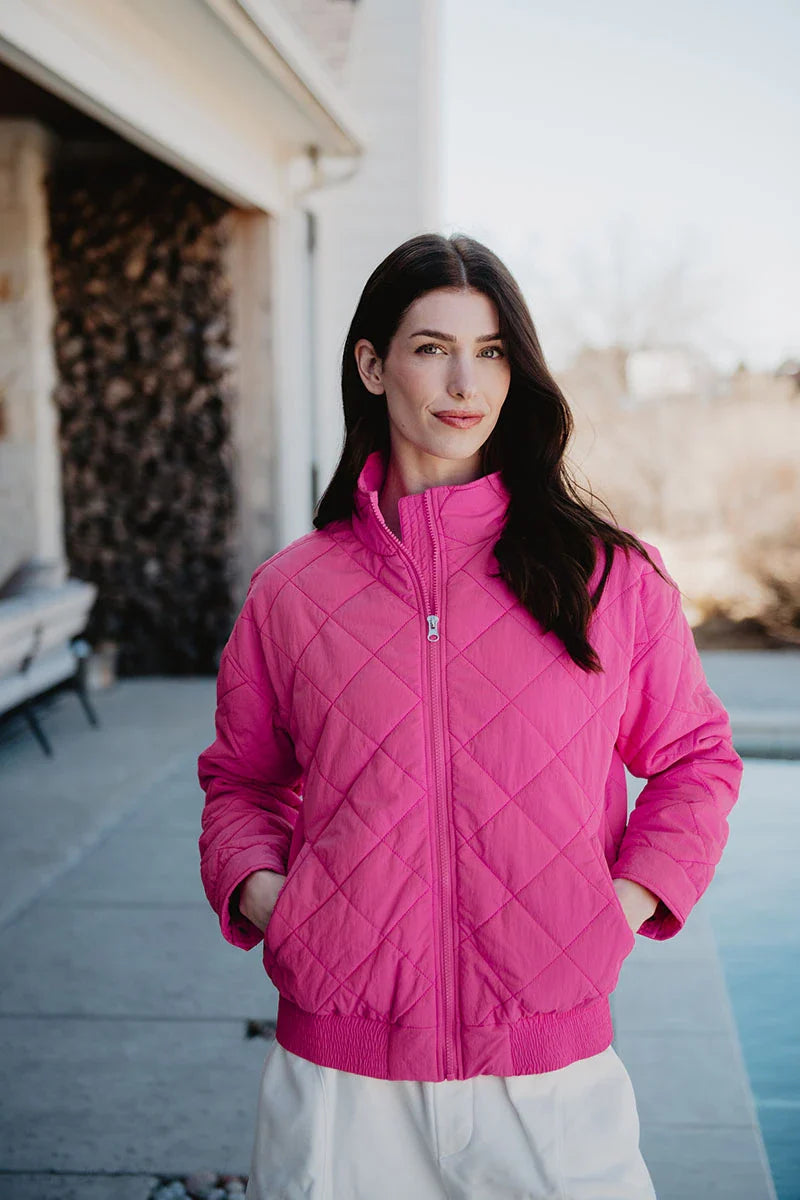 A woman with long dark hair stands outdoors in the Katydid Hot Pink Sporty Diamond Quilted Jacket and white pants, hands in her pockets. Behind her are a building, stacked firewood, and a blurred fall landscape.