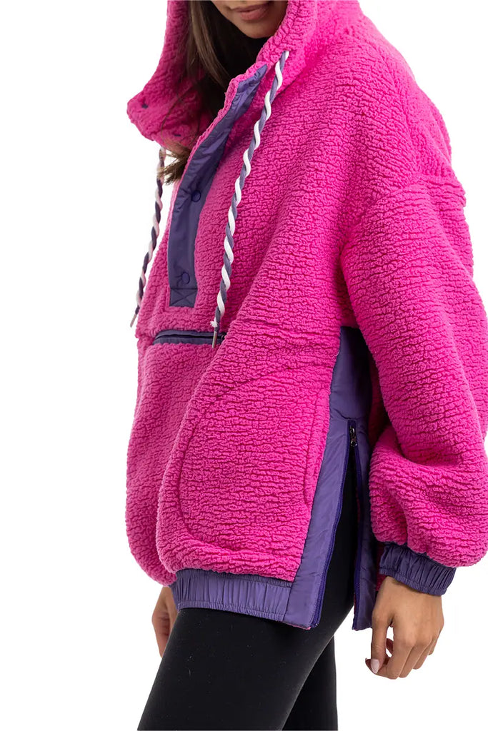 A person wears the Katydid Hot Pink Two-Toned Sherpa Pullover Jacket with purple accents, white-and-purple drawstrings, large pockets, and a side zipper, paired with black leggings. Their face is partially out of frame.