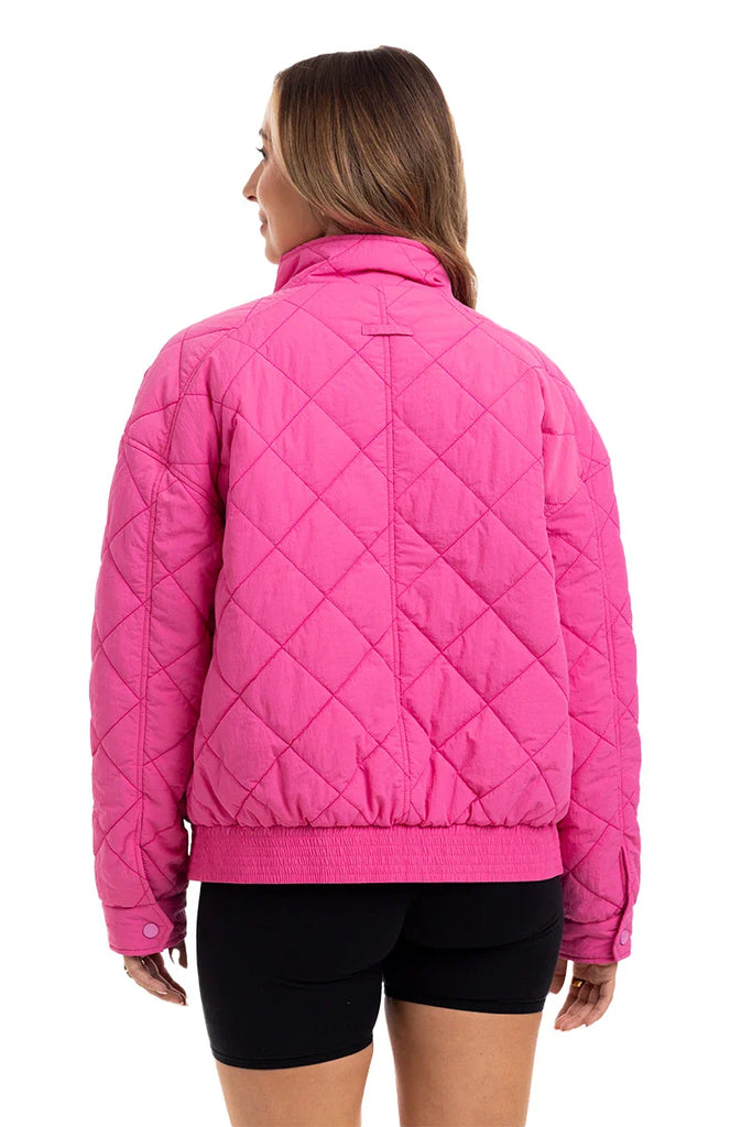 A woman, seen from behind, wears the Katydid Hot Pink Sporty Diamond Quilted Jacket with a high collar and black shorts. This lightweight jacket features a diamond pattern and elastic at the cuffs and waistband.