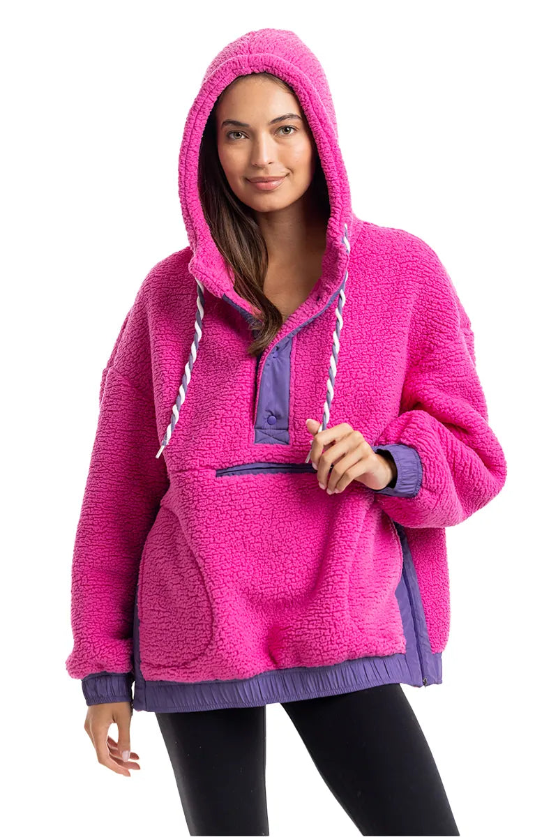 A woman smiles at the camera in the Katydid Hot Pink Two-Toned Sherpa Pullover Jacket, featuring a purple front pocket, trim, and hood, paired with black leggings.