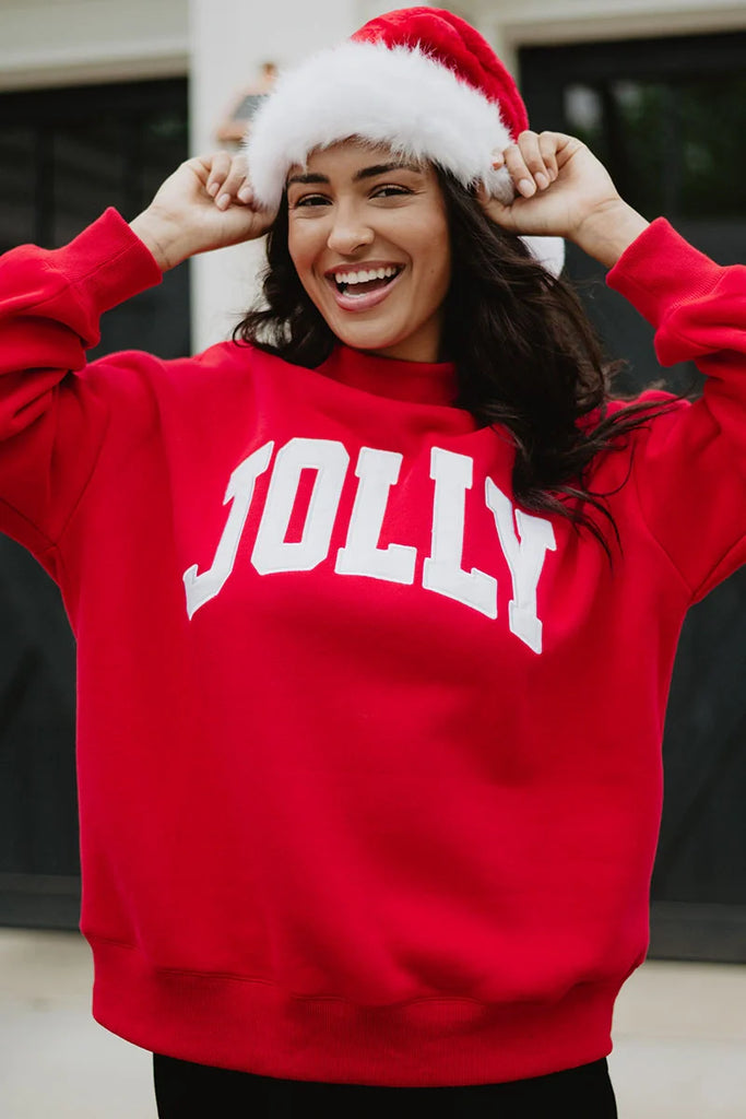 A smiling person in a red Santa hat wears the Katydid JOLLY Mockneck Sweatshirt, a festive pullover with JOLLY in bold white letters, while standing outdoors.