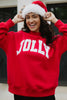 A smiling person in a red Santa hat wears the Katydid JOLLY Mockneck Sweatshirt, a festive pullover with JOLLY in bold white letters, while standing outdoors.