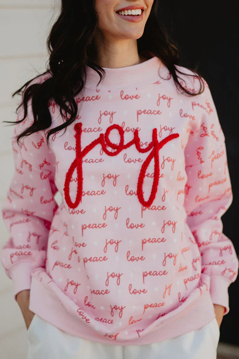 A woman wears the Katydid Joy Knit Mockneck Sweatshirt, a light pink holiday pullover for women with red and pink “joy,” “love,” and “peace” prints all over, plus joy boldly stitched in large red letters across the chest.