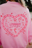 A person wears the Katydid Literally Just A Girl Mockneck Pullover, a cute pink sweatshirt with a large red heart on the back featuring embroidered shoes, bows, drinks, flowers, cake, and Literally Just A Girl in the center.