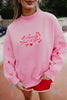 A woman wears the Katydid Literally Just A Girl Mockneck Pullover in light pink with red doodles and text, paired with red shorts with white trim, while standing on a tiled floor.