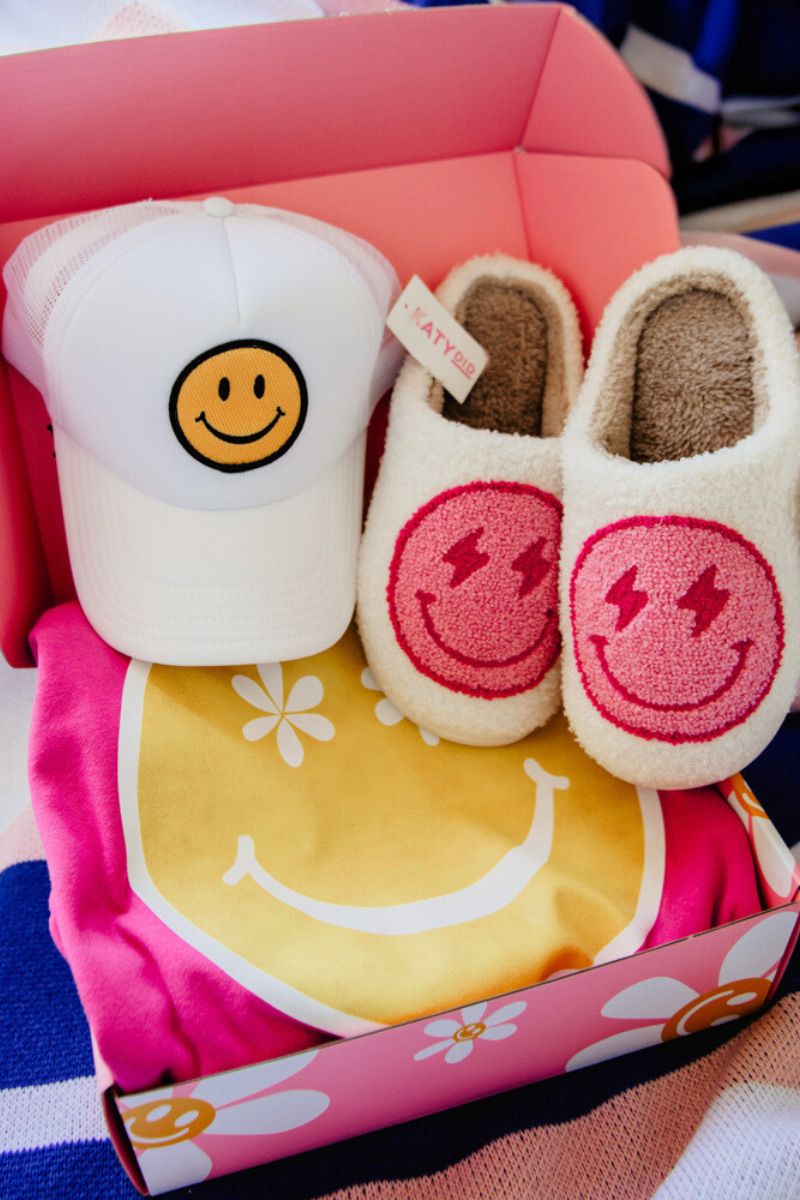 A pink gift box contains a white cap and slippers with yellow smiley faces. A yellow and pink shirt with a flower pattern lies beneath.