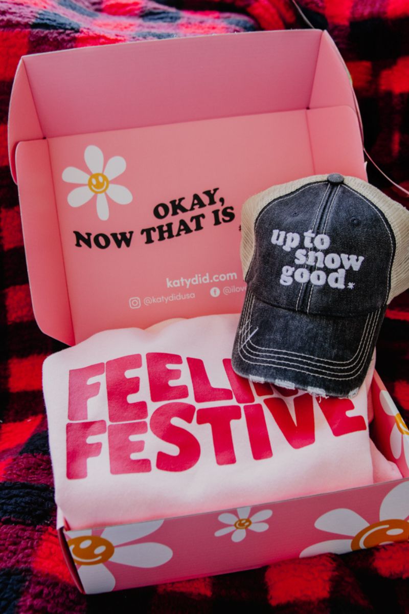 Open pink box with a daisy design, displaying a black cap reading "up to snow good" and a white shirt with "FEELING FESTIVE" in bold red.