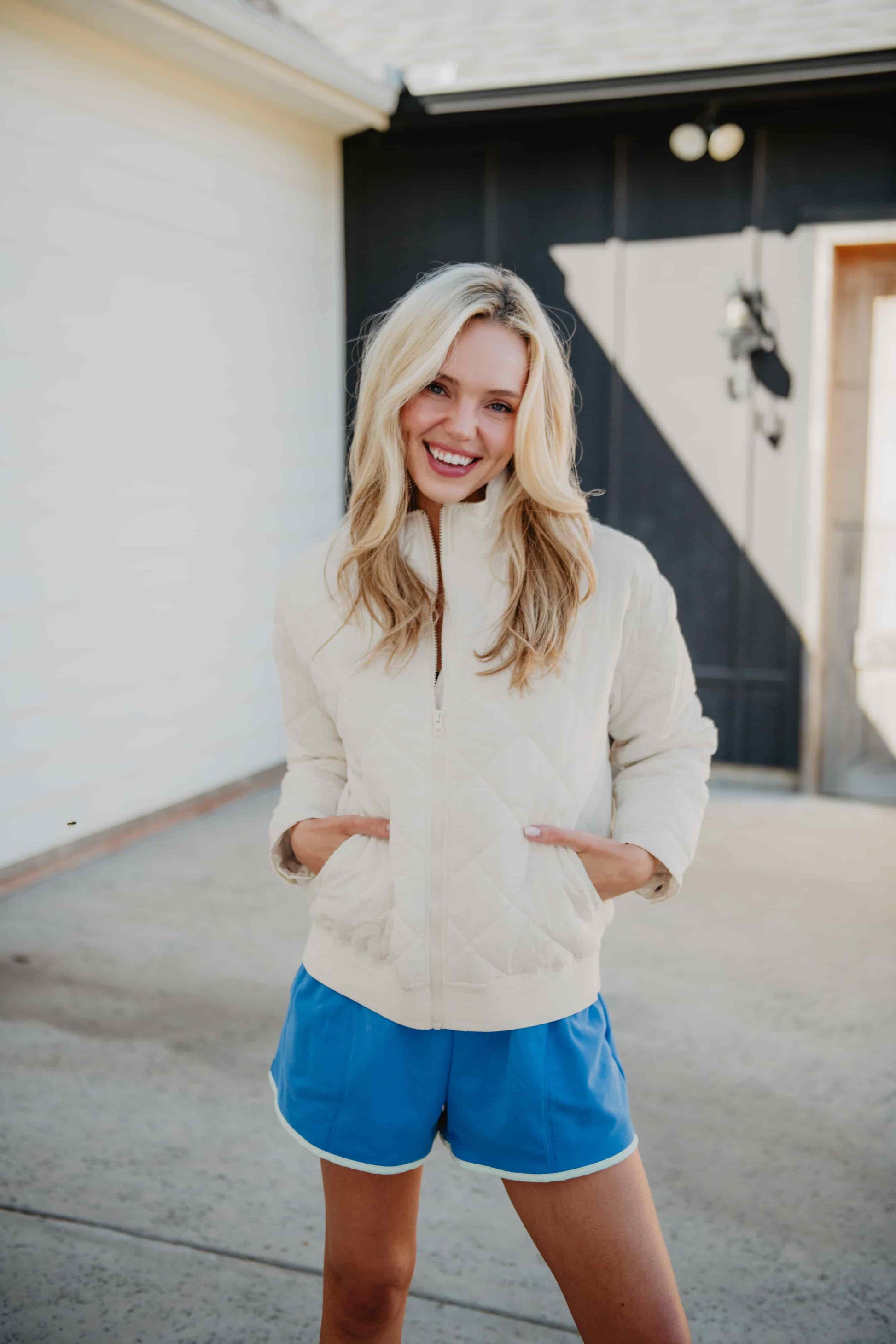 A smiling woman with long blonde hair stands outside a building, hands in her pockets, wearing the Katydid Cloud White Sporty Diamond Quilted Jacket and blue athletic shorts.