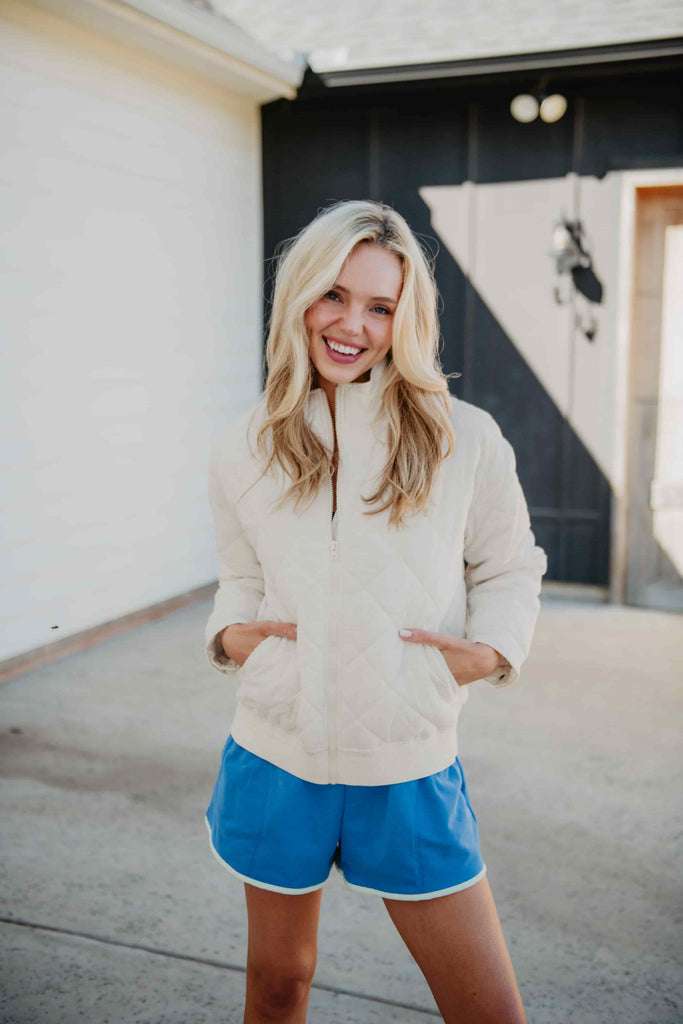 A smiling woman with long blonde hair stands outside a building, hands in her pockets, wearing the Katydid Cloud White Sporty Diamond Quilted Jacket and blue athletic shorts.