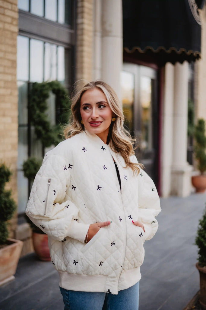 A woman with blonde hair stands outdoors before a building, smiling slightly with hands in her pockets, wearing the Katydid Cream and Black Bows Oversized Quilted Jacket.