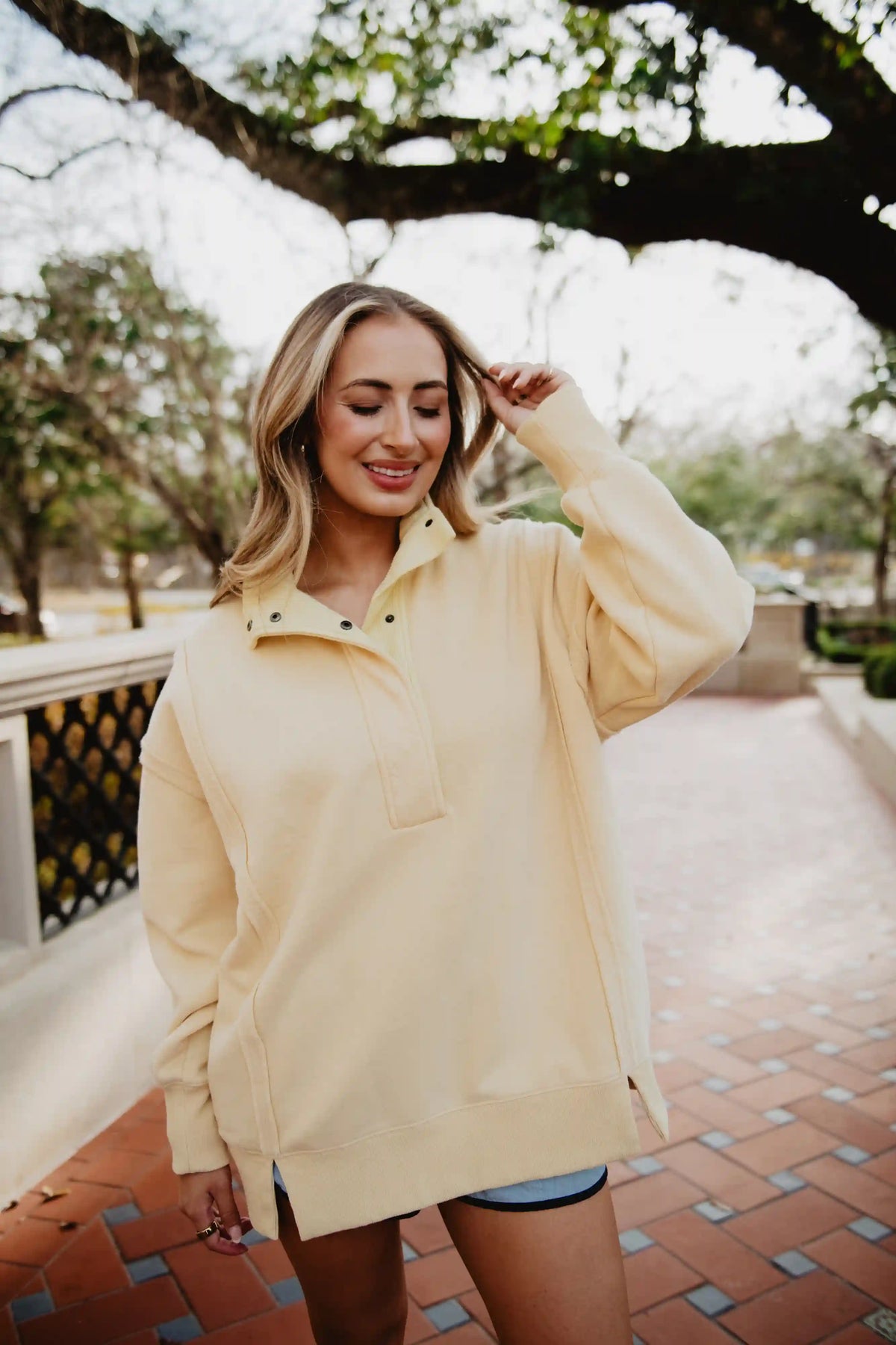 A woman with blonde hair smiles while walking outdoors in a Katydid Yellow Vintage Wash Pullover Sweatshirt and denim shorts, surrounded by trees and greenery.