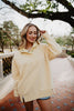 A woman with blonde hair smiles while walking outdoors in a Katydid Yellow Vintage Wash Pullover Sweatshirt and denim shorts, surrounded by trees and greenery.