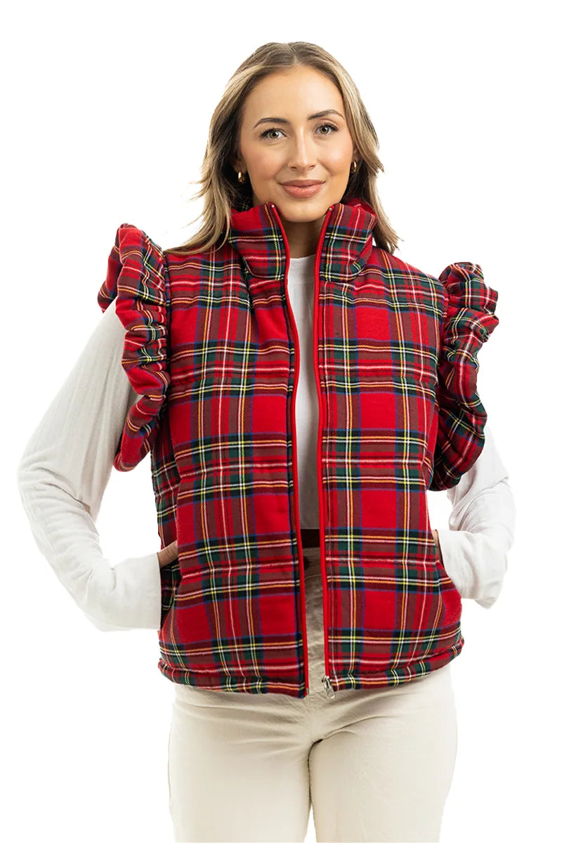 A woman smiles at the camera, hands in pockets, wearing the Katydid Royal Stewart Plaid Ruffle Puffer Vest over a white long-sleeve top and light pants—an ideal cheerful holiday look against a white background.