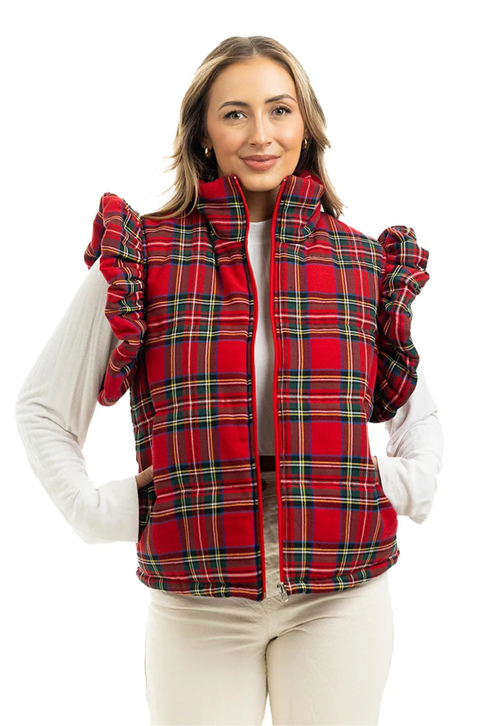A woman smiles at the camera, hands in pockets, wearing the Katydid Royal Stewart Plaid Ruffle Puffer Vest over a white long-sleeve top and light pants—an ideal cheerful holiday look against a white background.