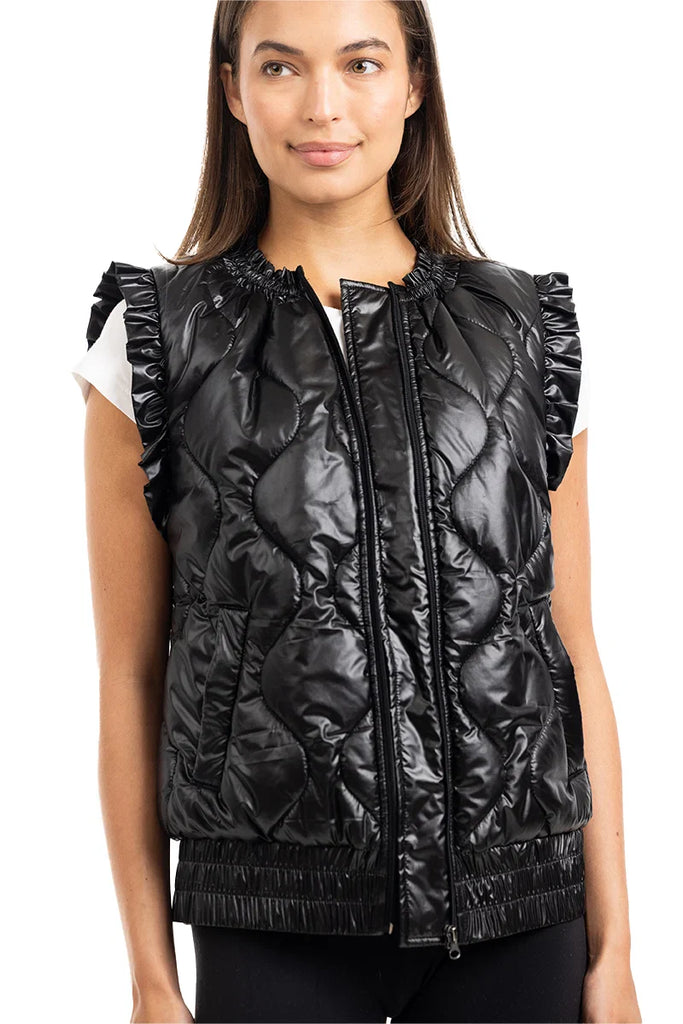 A woman wears the Katydid Black Metallic Quilted Vest with ruffled cap sleeves and a front zipper over a white short-sleeve top, standing and smiling—a stylish fall layer for women.