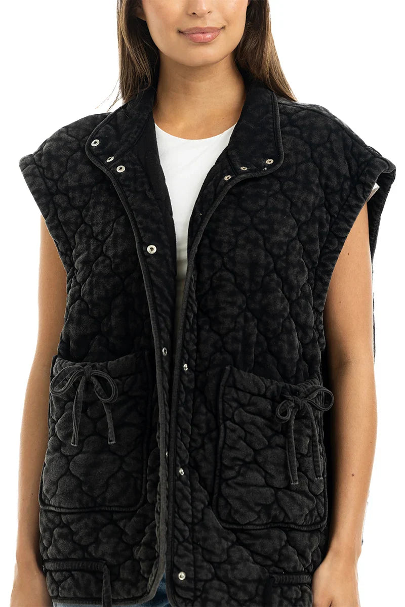 A woman wears the Katydid Charcoal Oversized Garment Washed Quilted Vest, featuring large pockets and a tie detail, over a white shirt while standing against a plain white background.