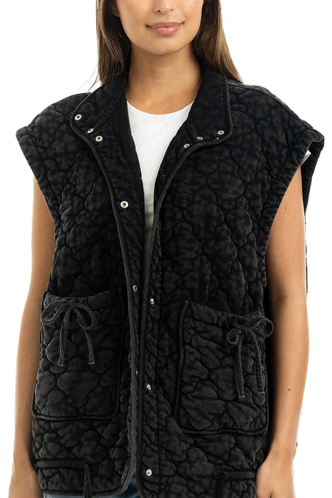 A woman wears the Katydid Charcoal Oversized Garment Washed Quilted Vest, featuring large pockets and a tie detail, over a white shirt while standing against a plain white background.
