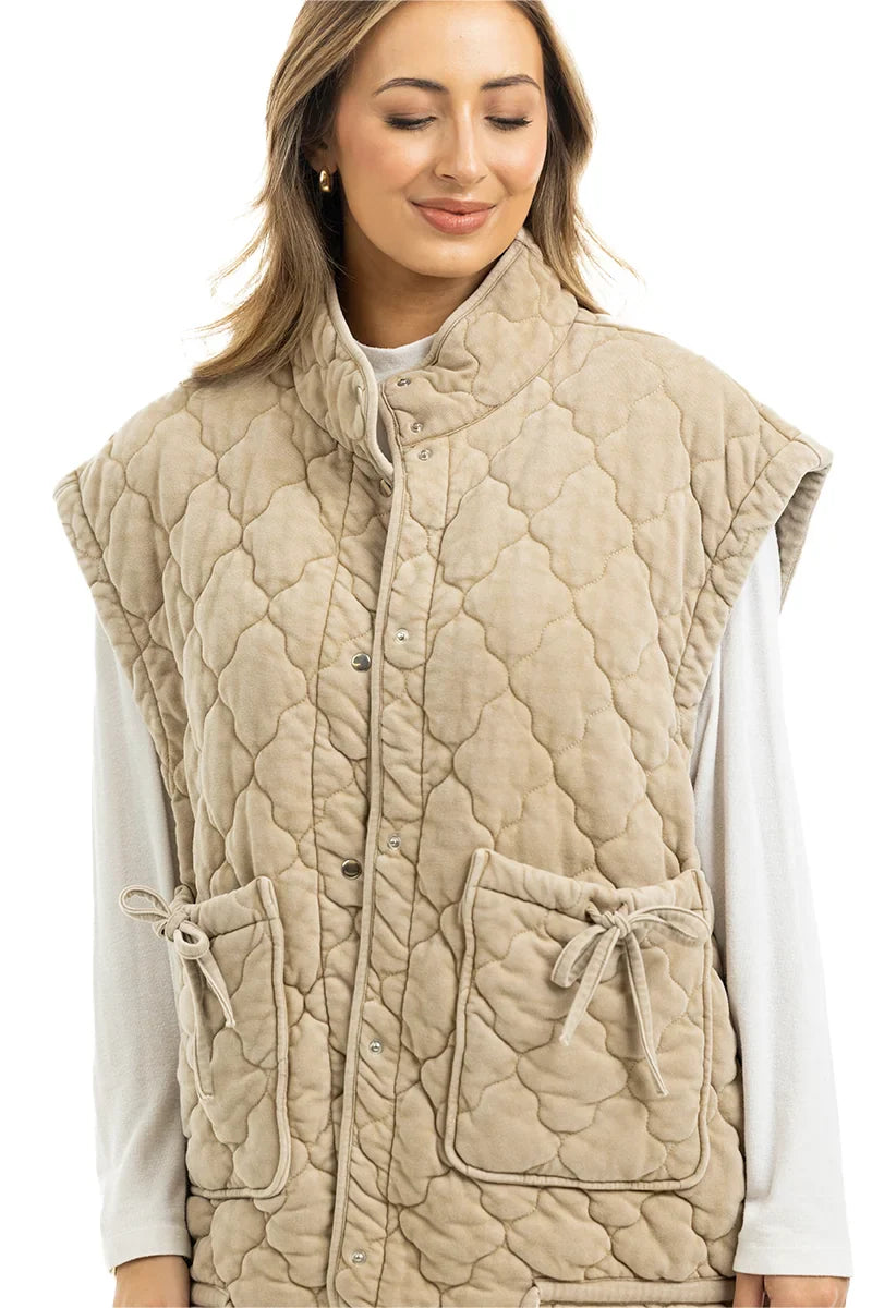 A woman with light brown hair smiles with her eyes closed, wearing the Katydid Beige Oversized Garment Washed Vest featuring snap buttons and two large front pockets tied with bows over a long-sleeve white top.