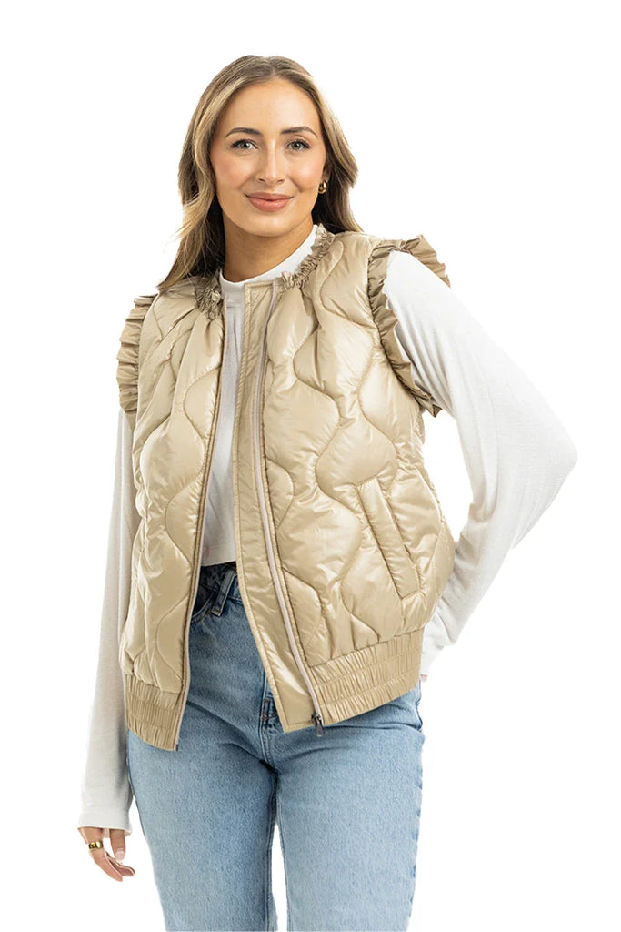 A woman in a Katydid Soft Tan Metallic Quilted Vest over a white long-sleeve shirt and blue jeans stands smiling against a white background.