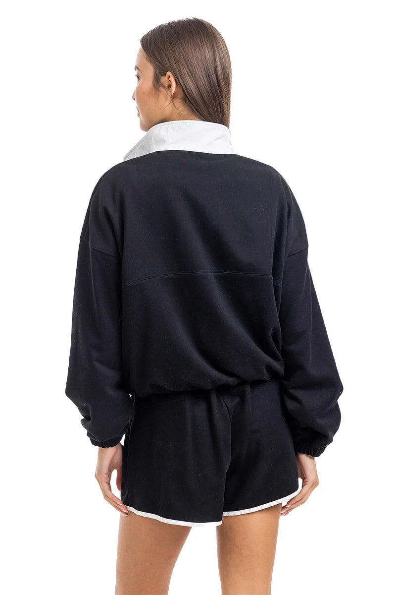 A woman is seen from the back in the Katydid Black  White Quarter Snap Athletic Set Womens, pairing the long-sleeve pullover with matching black shorts trimmed in white, her straight brown hair contrasting a plain white background.