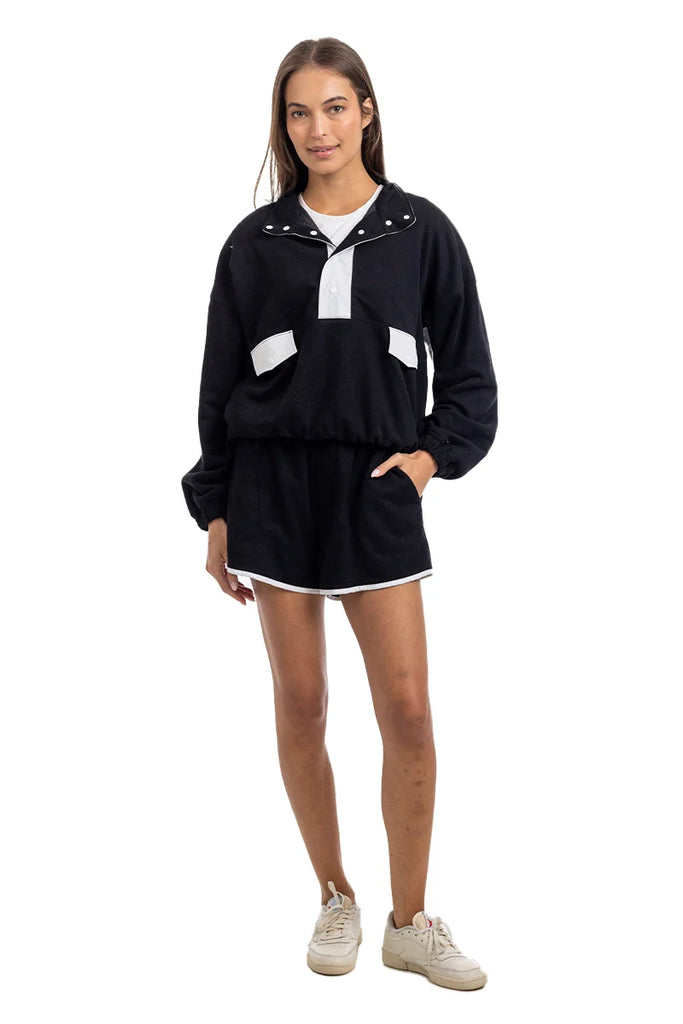 A woman poses against a white backdrop in the Katydid Black  White Quarter Snap Athletic Set for women, featuring a black long-sleeve pullover with white accents and matching black shorts trimmed in white, paired with white sneakers.