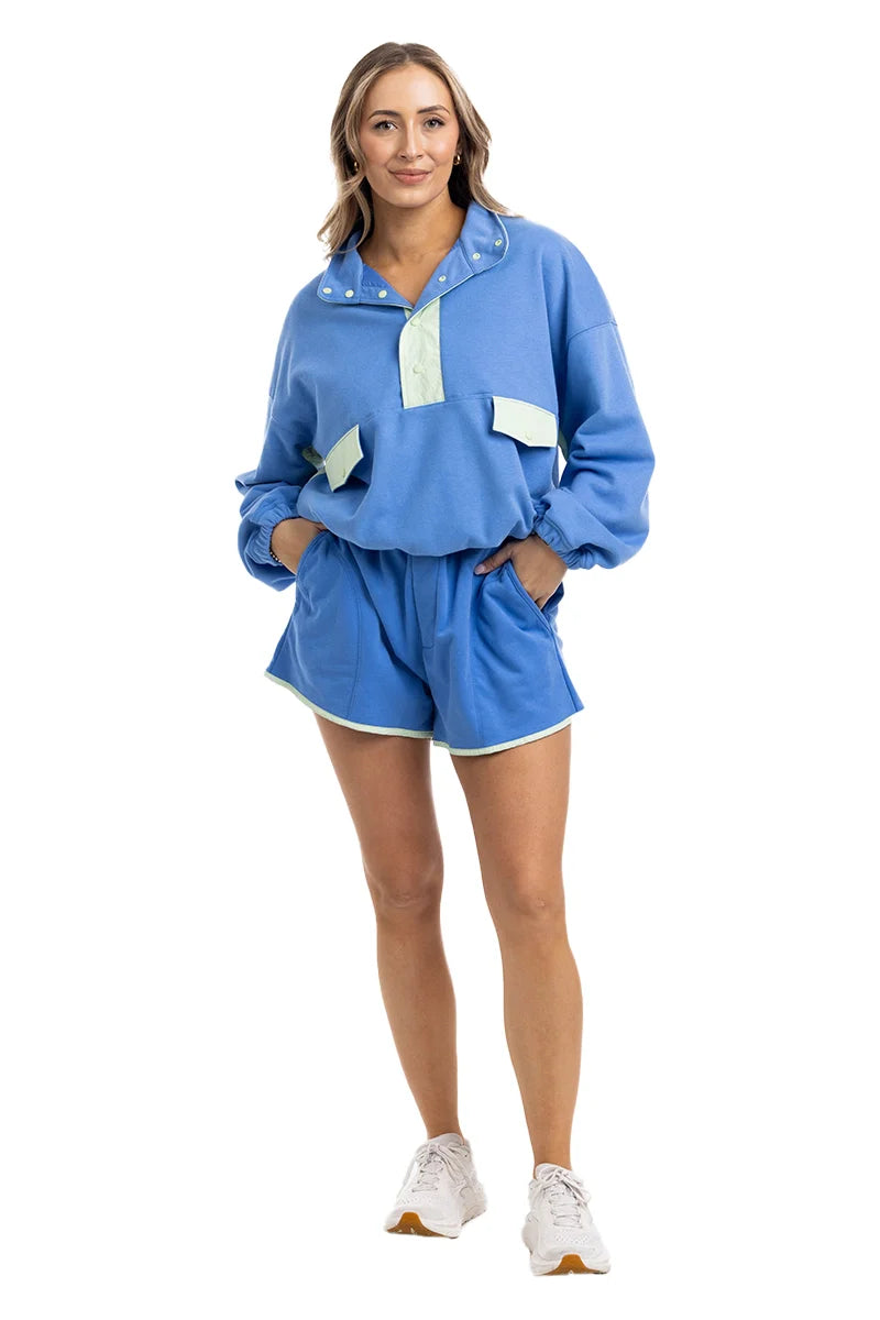 A woman smiles in the Katydid Blue  Green Quarter Snap Athletic Matching Set, featuring front-pocket top and matching shorts with white trim. She pairs it with white sneakers for an effortless athletic lounge look.