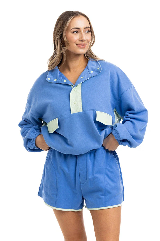 A woman smiles in the Katydid Blue  Green Quarter Snap Athletic Matching Set, featuring a light blue pullover with buttons and front pockets, paired with high-waisted green shorts, all set against a white background.