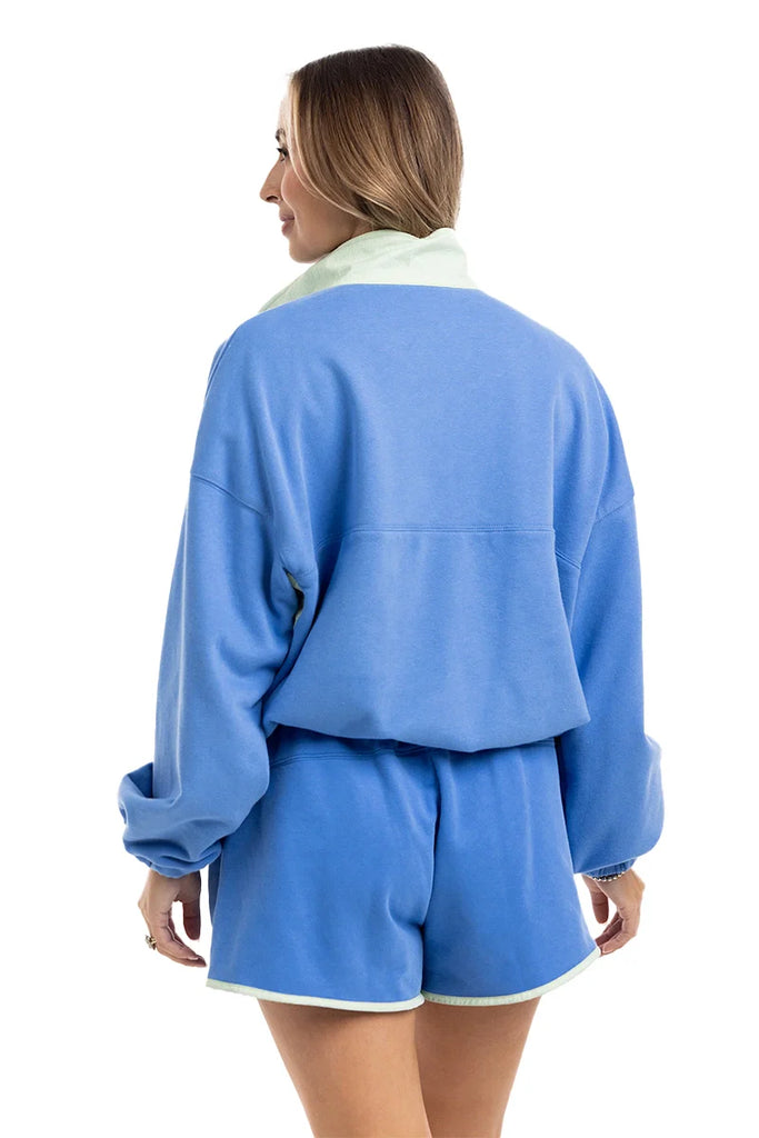 A woman with light brown hair, wearing rings, faces away from the camera in a Katydid Blue  Green Quarter Snap Athletic Matching Set—an athletic fit lounge set with a long-sleeve top and matching shorts.
