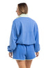 A woman with light brown hair, wearing rings, faces away from the camera in a Katydid Blue  Green Quarter Snap Athletic Matching Set—an athletic fit lounge set with a long-sleeve top and matching shorts.